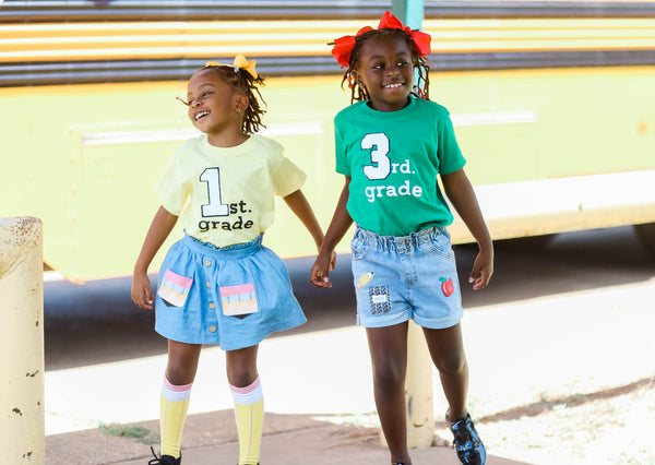 little girls wearing a yellow and green first grade and third grade shirts with custom patch numbers and a screen print of grade. Cute black girl magic photo behind a school bus.
